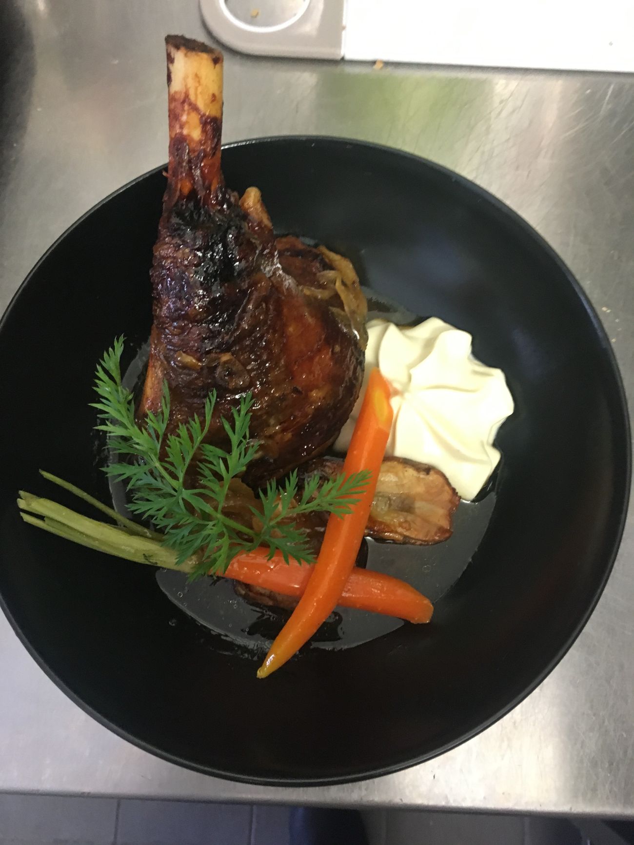 Un jarret d'agneau laqué dans un bol noir, avec carotte, crème et fanes